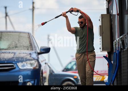 Confident man washing his car on self car-washing station. Cleaning car using active foam. Young man cleaning his auto by water gun. Stock Photo