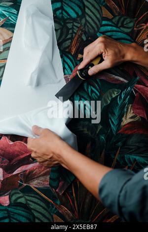 a caucasian man, wearing a gray working coat, is using a paint scraper to remove a strip of floral patterned wallpaper from a wall Stock Photo