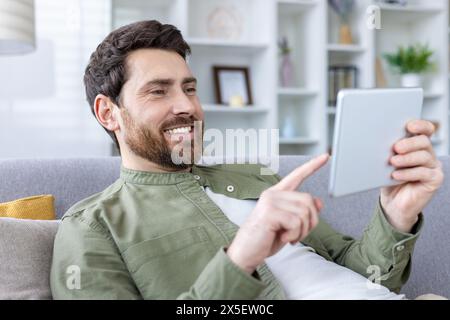 A cheerful, bearded man comfortably using a digital tablet while sitting on a couch. His relaxed posture and smile suggest he is enjoying his activity. Stock Photo