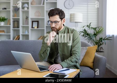 A thoughtful middle-aged man works on his laptop from a well-decorated living room. His expression is one of deep concentration, indicative of professional dedication or problem-solving. Stock Photo