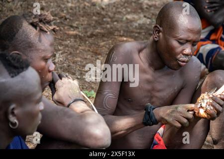 Members of Mursi Tribe in Ethiopia, in Omo valley, gathered around fire, eating meat from animal bones, primitive life in remote place in Africa Stock Photo