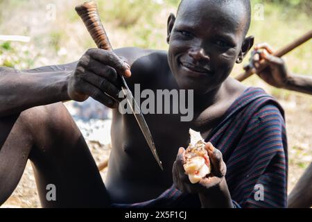 Members of Mursi Tribe in Ethiopia, in Omo valley, gathered around fire, eating meat from animal bones, primitive life in remote place in Africa Stock Photo