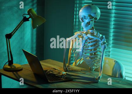 Waiting concept. Human skeleton sitting at wooden table with laptop indoors Stock Photo