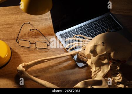 Waiting concept. Human skeleton sleeping at wooden table with laptop Stock Photo