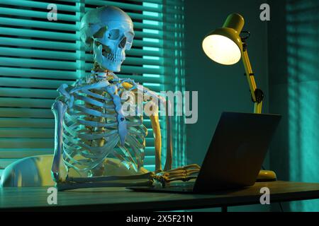 Waiting concept. Human skeleton sitting at wooden table with laptop indoors Stock Photo