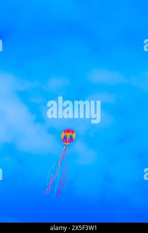 Colorful Kite Flying High in a Bright Blue Sky with Light Clouds Stock Photo