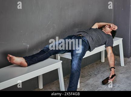 Depression, poverty and drunk man sleeping with beer on bench outdoor for addiction, disaster or mental health crisis on wall background. Homeless Stock Photo