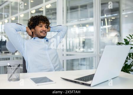 A young businessman relaxes at his desk with hands behind his head, looking thoughtful. The modern office setting with bright lighting encapsulates a moment of calm and contemplation during a busy workday. Stock Photo