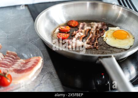 A pan of scrambled eggs, bacon and cherry tomatoes sits on the stove in the kitchen next to a plate of fresh tomatoes and raw bacon. High quality phot Stock Photo