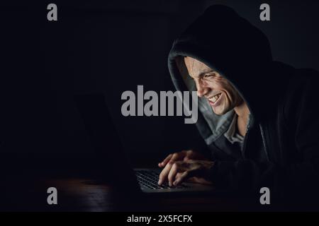 Dark, basement and man at laptop for hacking, online programming and cybersecurity at night. Software, developer or hacker in hoodie coding for Stock Photo