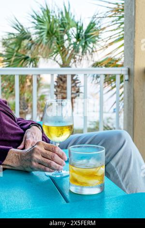 Woman seated at small table on balcony with a glass of white wine and a glass of bourbon on ice or on the rocks. Beach life in Destin Florida, USA. Stock Photo