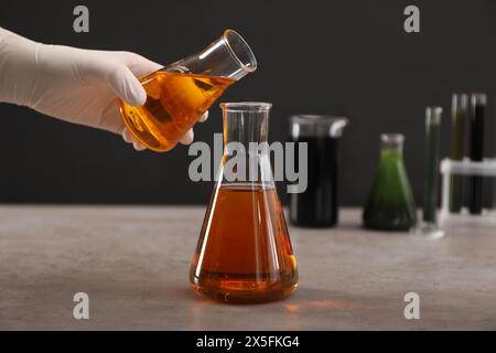 Woman pouring yellow crude oil into flask at grey table against dark background, closeup Stock Photo