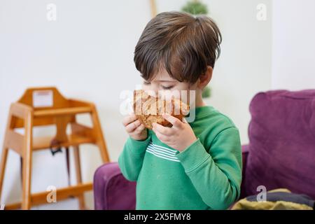 cute young boy in a green sweater eating a croissant on the couch in a cafe Stock Photo