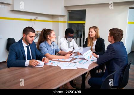 Multicultural team led by female boss analyzes graphs in office boardroom. Group engages in discussion at corporate meeting. Mixed-race professionals Stock Photo