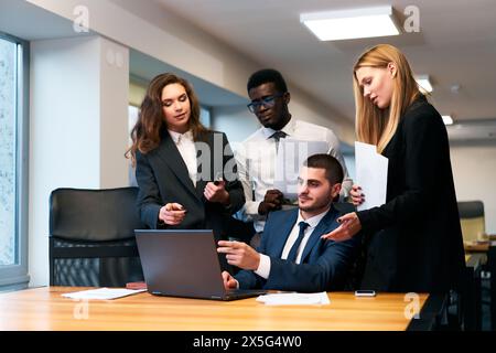 Multiethnic business team collaborates in office space, working on project with laptop. African and Caucasian men, women discuss strategies. Pro group Stock Photo