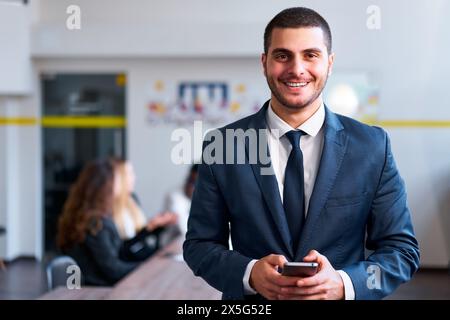 Smartly dressed eastern businessman smiles, holding smartphone in modern office. Colleagues work in background. Pro uses fintech app for efficient Stock Photo