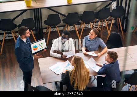 Arabic executive shares data on tablet with multiethnic team in modern office. Colleagues discuss strategy, graphs. Engaged diverse group analyzes Stock Photo