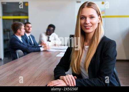 Confident blonde businesswoman leads meeting at modern office. Young caucasian female boss sits at table with multiethnic team. Colleagues discuss Stock Photo