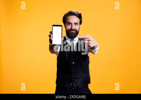 Confident waiter points to white screen on camera, presenting isolated mockup blank display on mobile phone. Young butler with suit and tie shows empty device with copyspace template. Stock Photo