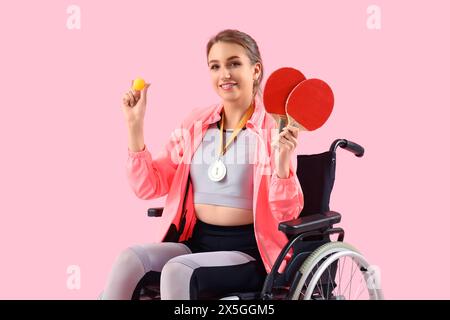 Sporty young woman in wheelchair with first place medal and ping pong rackets on pink background Stock Photo