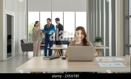 Young asian woman working with laptop computer on wooden table. In the background, three other individuals are engaged in a conversation, sharing a do Stock Photo