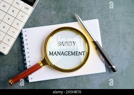 Safety Management text written through a magnifying glass on a notebook in a composition with a calculator, a pen on a gray background Stock Photo