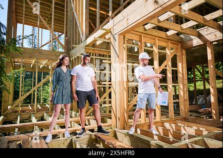 Man designer with plan in hand shows to couple the process of constructing wooden framed house. Investors inspecting their future dwelling near forest. Concept of contemporary ecological construction. Stock Photo