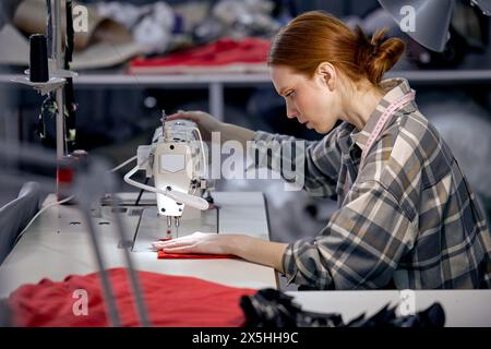 seamstress lady in casual wear sit behind table using sewing machine, making modern clothes, young redhead caucasian woman at workshop, holding red fa Stock Photo
