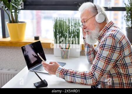 Handsome elderlycaucasian man sitting alone, using modern laptop and headset, bearded male having video call on smartphone, attending video conference or webinar, use modern technologies Stock Photo