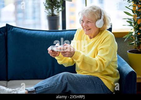 funny european senior woman with gray hair with joystick play online game at spare time in room interior, alone. Elderly people and modern gadget, retired people and modern technologies concept Stock Photo