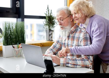 aged spouses using apps on laptop. Happy senior couple having fun online, surfing the Net. Senior people active social networks users spend time together, watching something interesting Stock Photo