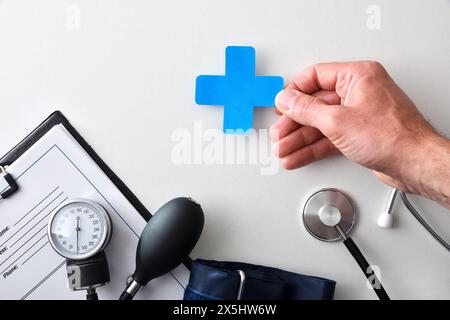 Concept of medical control with hand holding cutout of health cross on white table with blood pressure monitor stethoscope and folder with history. To Stock Photo