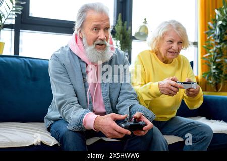 elderly couple playing video game console with joysticks sitting on couch in living room. Playful people family having fun spend leisure time together. gray hair man and woman enjoy entertaining Stock Photo