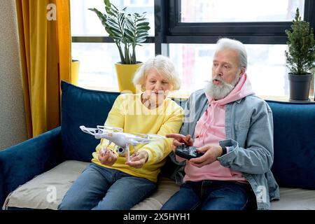Happy elderly caucasian couple launch a drone at home, man holding console in hands. caucasian retired spouses enjoy using modern technologies, learning to use at home in living room Stock Photo