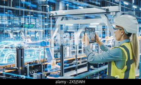 Futuristic Car Factory Digitalization Industry 4.0: Female Automotive Engineer Using Augmented Reality Tablet Computer to Scan Automated Robot Arm Assembly Line Manufacturing Electric Vehicles Stock Photo