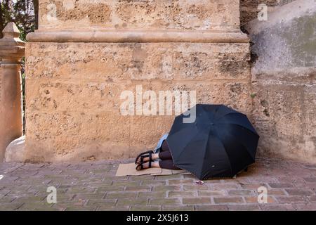 A homeless woman sleeps on the floor at the entrance to the cathedral in Malaga, Spain, covered by a black umbrella Stock Photo