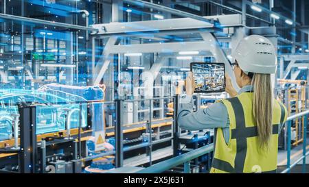 Futuristic Car Factory Digitalization Industry 4.0: Female Automotive Engineer Using Augmented Reality Tablet Computer to Scan Automated Robot Arm Assembly Line Manufacturing Electric Vehicles Stock Photo