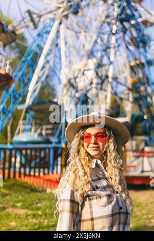 A woman wearing a hat and sunglasses stands in front of a Ferris wheel Stock Photo