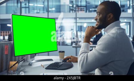 Diverse Modern Office: Handsome Black IT Technician Using Desktop Computer with Green Chroma Key Screen. Engineer Administrator Work on e-Commerce Development, Project Marketing, Big Data Analysis Stock Photo