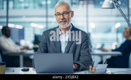 Modern Office: Portrait of Successful Middle Aged Bearded Corporate Businessman Working on a Laptop at his Desk, Smiling. Diverse Workplace with Professionals. Stock Photo