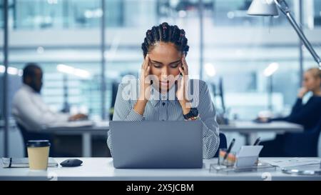 Young Black Woman Office Worker Uses Laptop, Feels Sudden Burst of Pain, Headache, Migraine. Overworked Accountant Feeling Project Pressure, Stress, Massages Her Head, Temples. Front View Portrait Stock Photo
