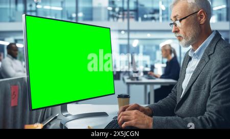 Handsome Senior Specialist Working on Desktop Computer with Green Chroma Key Screen Display in a Busy Corporate Office. Mature Male Manager Creatively Working In Multi-Ethnic International Workplace Stock Photo