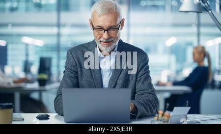 Modern Office: Portrait of Successful Middle Aged Bearded Corporate Businessman Working on a Laptop at his Desk, Smiling. Diverse Workplace with Professionals. Stock Photo