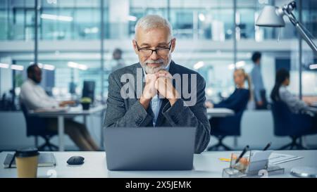 Modern Office: Portrait of Successful Middle Aged Bearded Businessman Working on a Laptop at his Desk. Smiling Corporate Worker. Multi-Ethnic Workplace with Happy Professionals. Front View Shot Stock Photo