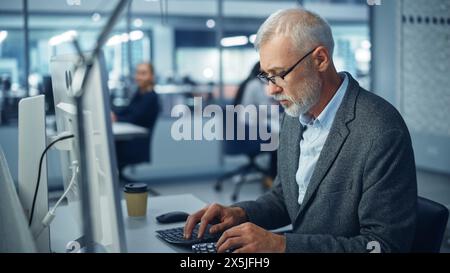Modern Office: Portrait of Successful Middle Aged Bearded Businessman Working on a Laptop at his Desk. Smiling Corporate Worker. Multi-Ethnic Workplace with Happy Professionals. Stock Photo