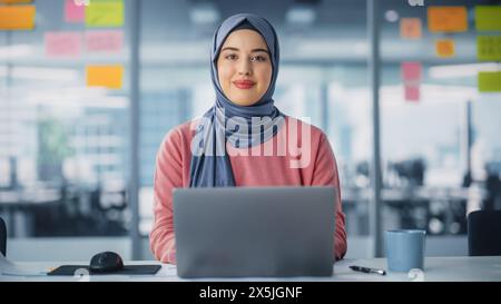 Modern Office: Portrait of Young Muslim Businesswoman Wearing Hijab Works on Laptop, Does Data Analysis, Looks at Camera and Smiles. Empowered Digital Entrepreneur Works on e-Commerce Startup Project Stock Photo