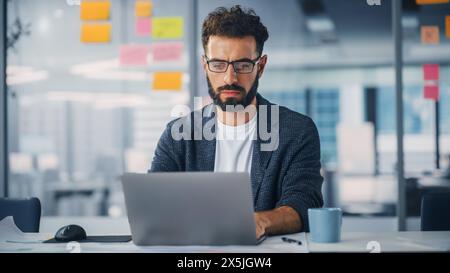Modern Office: Portrait of Young Stylish Businessman Wearing Glasses Works on Laptop, Does Data Analysis, Website Design, Creative Developemnt. Digital Entrepreneur Works on e-Commerce Startup Project Stock Photo