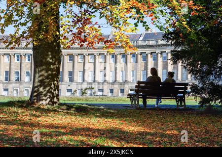 Two women bask in the autumn sunshine, sitting peacefully in front of Bath's iconic Royal Crescent, surrounded by vibrant autumn foliage. Stock Photo