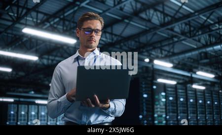 Male IT Engineer Working with Laptop in Data Center. Technician or Developer Standing at the Server Rack Corridor with a Laptop Computer. High-Speed Data Transfer, Analytics, Statistics Concept Stock Photo