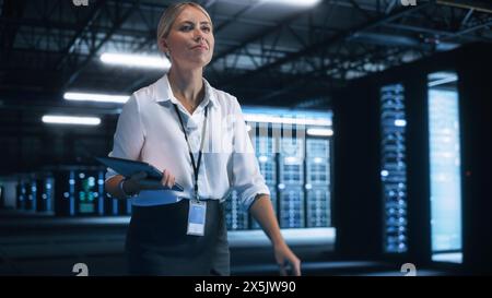 Young Caucasian Female Employee Uses Tablet Computer in System Control Monitoring Center. In the Background Night Corridor of her Office. Smooth Server Operation, Analytics, Statistics Concept Stock Photo